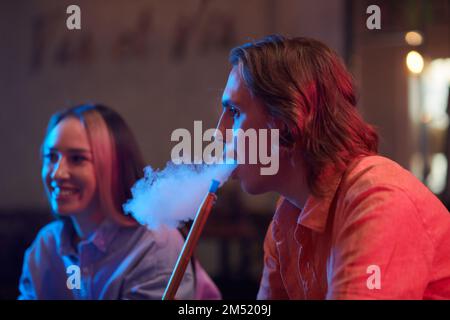 Side view of young man smoking hookah when gathering with friends in lounge bar Stock Photo