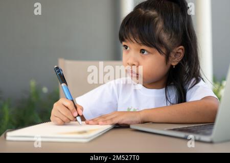Asian girl student holding a pen for taking notes and doing homework and using the laptop for online learning class at home, Education and distance le Stock Photo