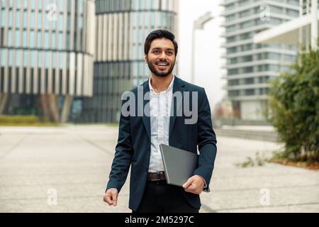 Smiling indian businessman in suit with laptop near office building Stock Photo