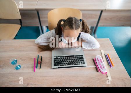 Caucasian girl sits at a desk at school and studies at a laptop. Hourglass on the table. Stock Photo
