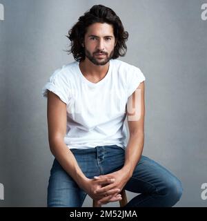Hes a real catch. Studio shot of a handsome young man sitting on a stool against a grey background. Stock Photo