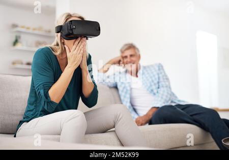 She cant believe its not real. a mature woman using a VR headset with her husband in the background. Stock Photo