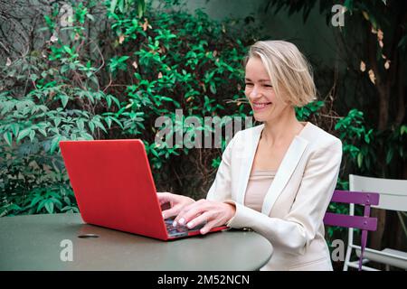 Businesswoman smiling while working with a laptop sitting on a table outdoors. Stock Photo