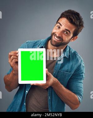 Well, well, well...what do we have here. Studio shot of a handsome young man showing a digital tablet with a green screen against a grey background. Stock Photo