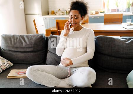 Young beautiful smiling happy pregnant afro woman sitting on sofa holding her belly and talking on phone in cozy room at home Stock Photo