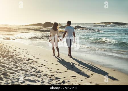 They enjoy long walks on the beach. Rearview shot of a carefree young couple walking hand in hand along the beach. Stock Photo