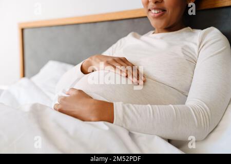 Adult beautiful pregnant smiling african woman holding her belly while liying on bed at home Stock Photo