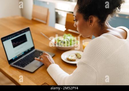 Back view of african woman looking on her laptop while eating salad and avocado toasts with glass of orange juice close-up in cozy kitchen at home Stock Photo