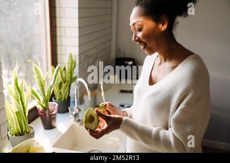 Adult beautiful african smiling pregnant woman peeling avocado in cozy kitchen at home Stock Photo
