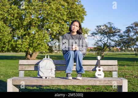 Girl with sad face, sitting on bench with smartphone and ukulele, looking upset and disappointed, being alone in park Stock Photo