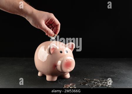 A person puts a coin in a piggy bank with a stack of coins next to it on a concrete table on a dark black background with copy space, using it as a Stock Photo