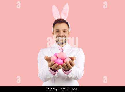 Smiling man wearing bunny ears holding Easter eggs isolated on pastel pink background. Stock Photo