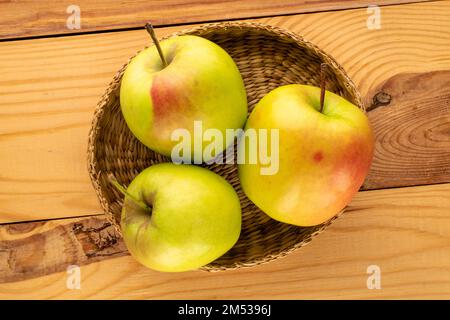 Three sweet green apples in a straw plate on a wooden table, macro, top view. Stock Photo