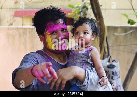 The Indian man holding a baby girl and smiling while pointing at the camera both covered in colorful paints Stock Photo