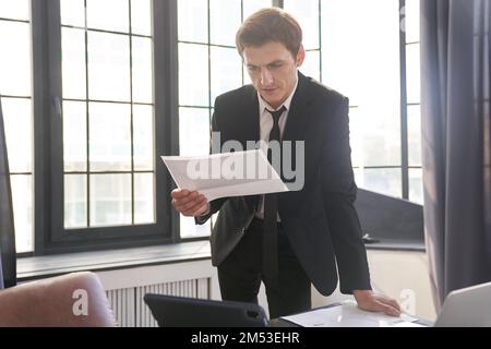 A businessman man examines a paper near the desk in the office against the background of a window. Business portrait of a businessman working in an office. Space for copying. High quality photo Stock Photo