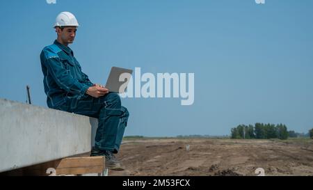 Caucasian male builder in hardhat sits on floor slabs and uses laptop at construction site.  Stock Photo