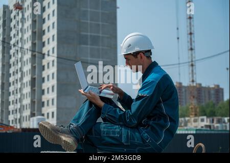 Caucasian male builder in hardhat sits on floor slabs and uses laptop at construction site.  Stock Photo