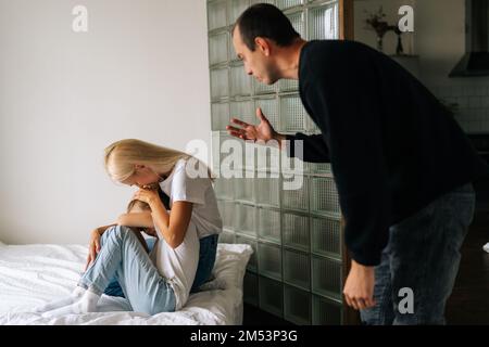 Back view of scared mother and daughter hugging each other sitting on sofa while aggressive standing father angrily yells at them at home. Stock Photo