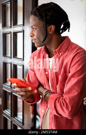 Thoughtful casual African American man with mobile phone stands indoors using messaging apps Stock Photo
