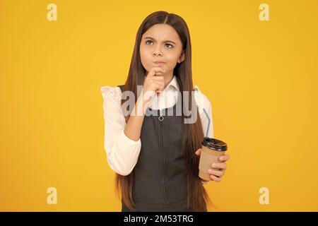 Thinking pensive clever teenager girl. Teenager child with coffee cup isolated on yellow studio background. Girl drinking take away beverage. Stock Photo