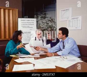 Businessmen and business women sitting around conference table discussing work issues papers on table, easel in background Stock Photo