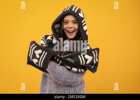 Warm hat with hood and scarf. Amazed teenager. Beautiful teen girl in a winter hat and a warm sweater. A child on a yellow isolated background Stock Photo