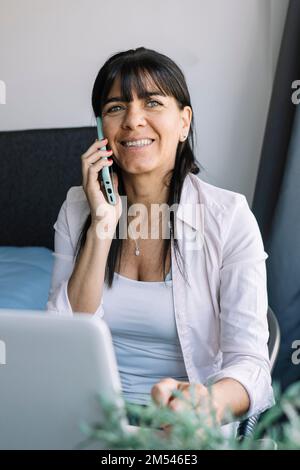 Smiling female entrepreneur using laptop while talking through smart phone at home office Stock Photo
