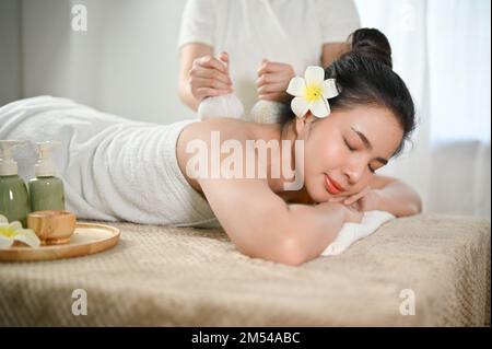 Beautiful and charming Asian woman relaxing in spa salon, getting body massage with spa herbal balls by a masseur. Thai spa salon concept Stock Photo