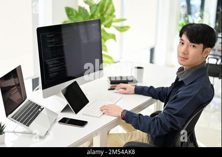 Professional and smart young Asian male programmer sits at his modern office desk, smiling at looking at the camera. Stock Photo