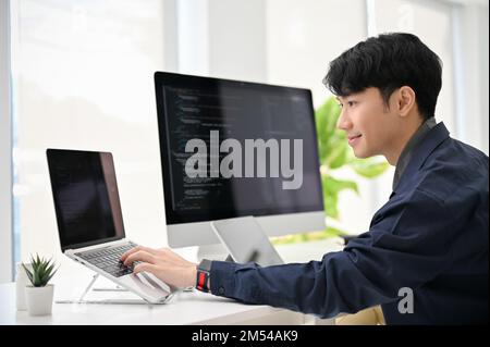 Professional and smart young Asian male programmer coding at his desk, working on his project, using laptop and computer. Stock Photo