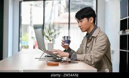 side view, Handsome and smart young Asian male programmer or freelance graphic designer remote working at the coffee shop, sipping coffee while using Stock Photo