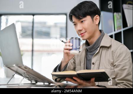 Smart and professional young Asian male programmer or web developer concentrating on his task, sipping coffee and using laptop computer. Stock Photo