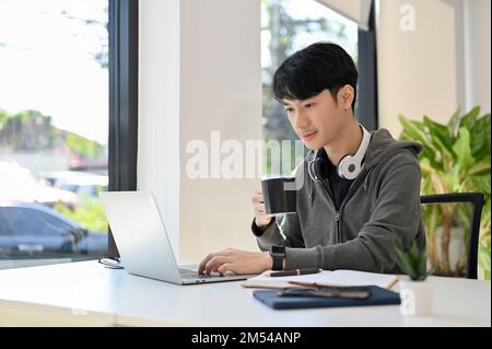 Smart young Asian male programmer or web developer sipping morning coffee while using laptop computer, working on his task. Stock Photo