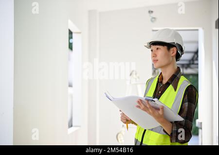 Handsome young Asian male engineer or architect wearing white hardhat, holding a building blueprint, inspecting the building at the construction site. Stock Photo