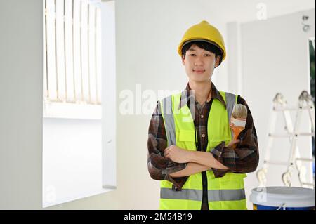 Smart and professional young Asian male construction worker in uniform and yellow hardhat stands with arms crossed, holding a paint brush, smiling and Stock Photo