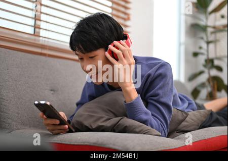 Relaxed and happy young Asian man lying on sofa, listening to music through his headphones and using his smartphone. Stock Photo
