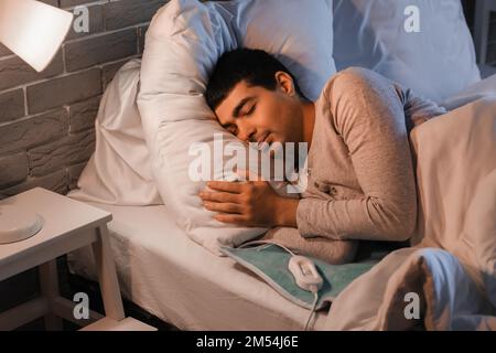 Young man sleeping on electric heating pad in bedroom at night Stock Photo