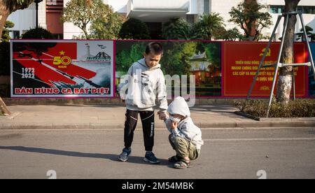 A young boy helps his baby brother near Communist party propaganda posters in front of the Hanoi People's Committee building in the centre of the Viet Stock Photo