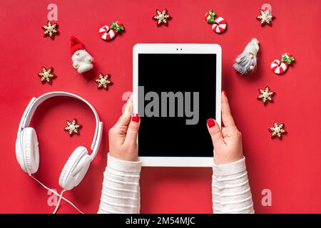 top view female hands holding tablet with black digital screen, Christmas decor, headphones on red table Flat lay Greeting electronic card, book, podcast, music Merry Christmas and Happy New Year. Stock Photo