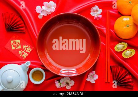Red plate with chopstick on red satin cloth background with tea set, ingots, oranges and red envelope packet or ang bao(word mean weath, lucky) for Ch Stock Photo