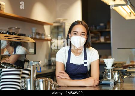 Covid-19 and cafe work. Asian girl barista in uniform, standing behind counter, works as bartender in coffee shop, wearing medical mask from Stock Photo