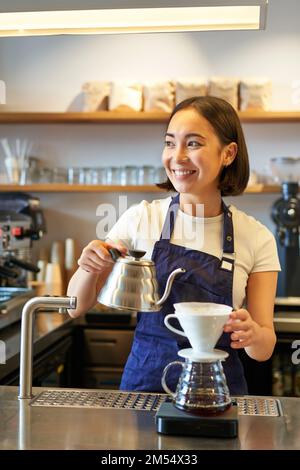 Vertical shot of smiling asian bartender, barista in blue apron, pouring water with small kettle, brewing coffee behind counter in her cafe Stock Photo