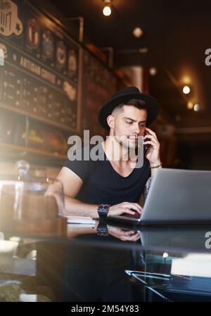 The hard work responsible for his cafes success. a young man using a phone and laptop while working in a coffee shop. Stock Photo