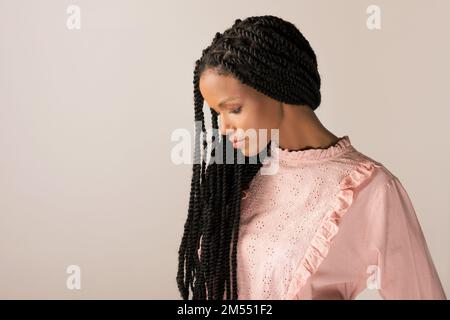 Young black female in pink retro blouse with long Afro braids looking down against gray background Stock Photo
