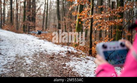 Rear view of a woman managing a drone floating in front of her with remote controller outside in winter forest. Focus on the drone. Stock Photo