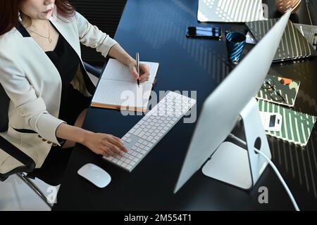 Overhead view of millennial businesswoman looking at computer screen and making notes on notebook Stock Photo