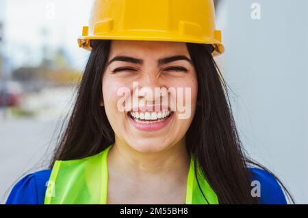 front view closeup portrait of happy young female engineer smiling with a laugh, standing on the street, wearing safety helmet and vest. she is lookin Stock Photo
