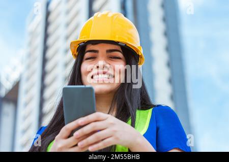 female engineer using the phone, smiling looking at the camera is standing on the street with buildings in the background Stock Photo