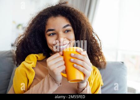 Cheerful smiling african american girl relaxing on couch with cup of coffee, resting at home, enjoying day off, wearing warm clothes Stock Photo