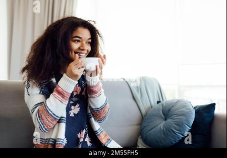 Cheerful smiling african american girl relaxing on couch with cup of coffee, resting at home, enjoying day off, wearing warm clothes Stock Photo